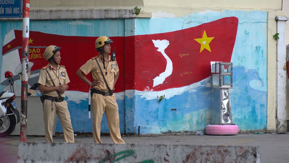 Polis berdiri di jalan di hadapan bendera Vietnam.